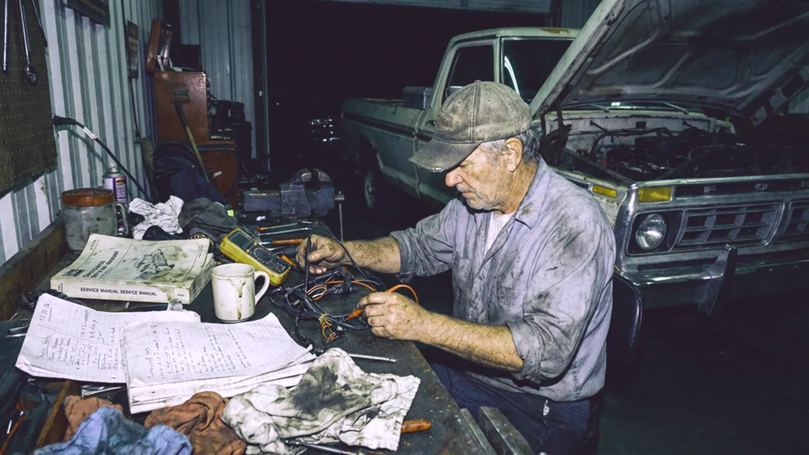Mechanic testing a wiring harness with a multimeter at a cluttered workbench, pickup truck with its hood up behind him
